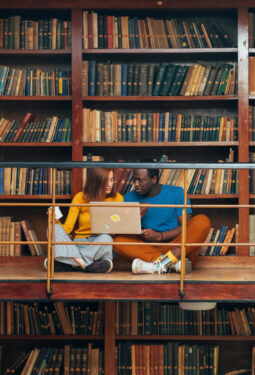 Male And Female Sharing A Laptop Infront Of A Library Shelf